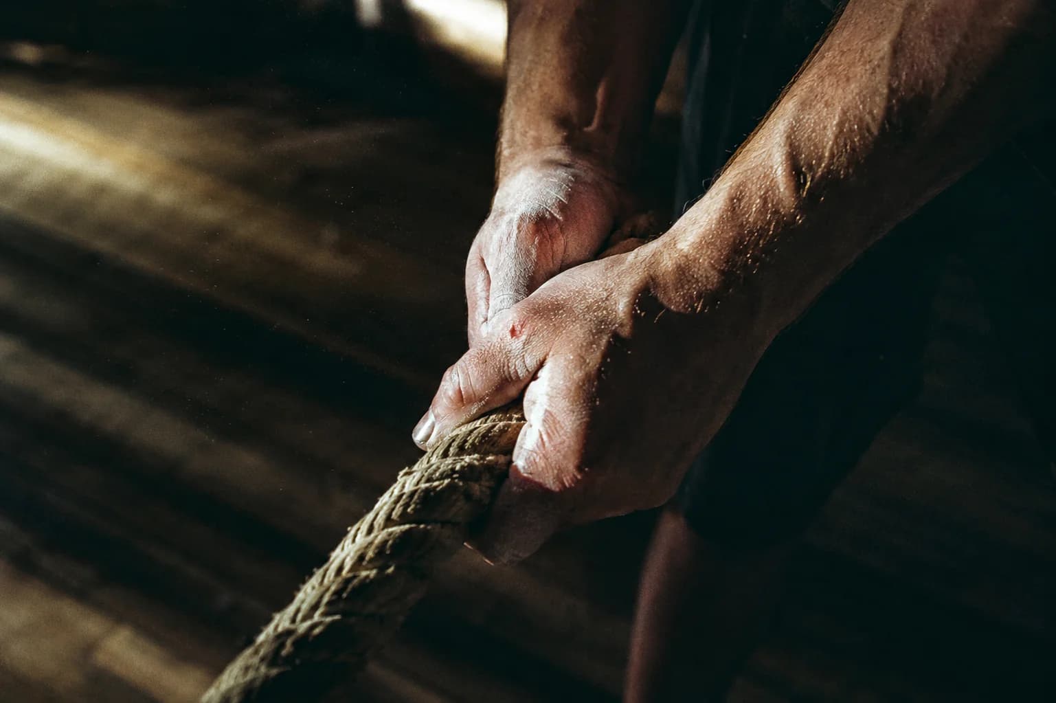 Chalk-dusted hands gripping a heavy rope mid-pull, hard side-light from a high window catching dust in the beam